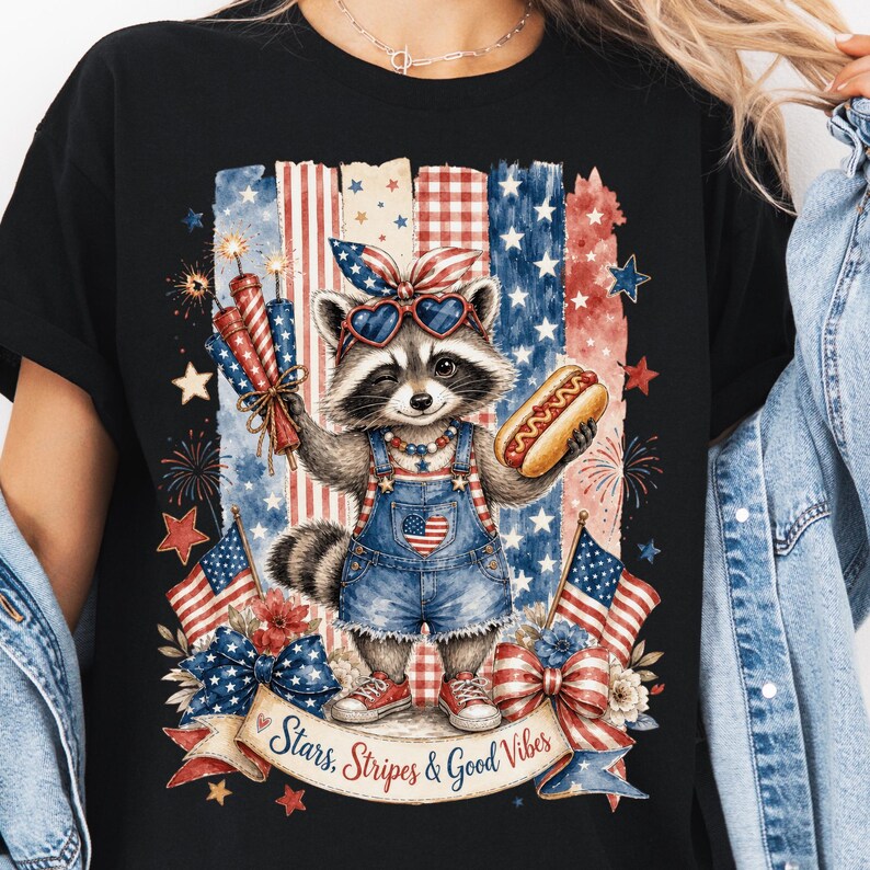 May include: Black t-shirt featuring a raccoon wearing heart-shaped sunglasses and denim overalls, holding fireworks and a hot dog. The design includes American flags, stars, and the text "Stars, Stripes & Good Vibes."