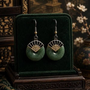 May include: A pair of jade earrings with a circular green stone design. The earrings feature a black and gold fan-shaped accent. The earrings are displayed on a dark green velvet surface inside a wooden box.