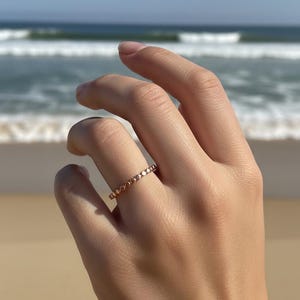 Puede incluir: Un anillo de oro rosa con un delicado dise&ntilde;o de cuentas. El anillo se muestra en una mano con un fondo de playa y oc&eacute;ano. El dise&ntilde;o del anillo presenta peque&ntilde;as cuentas espaciadas uniformemente alrededor de la banda.