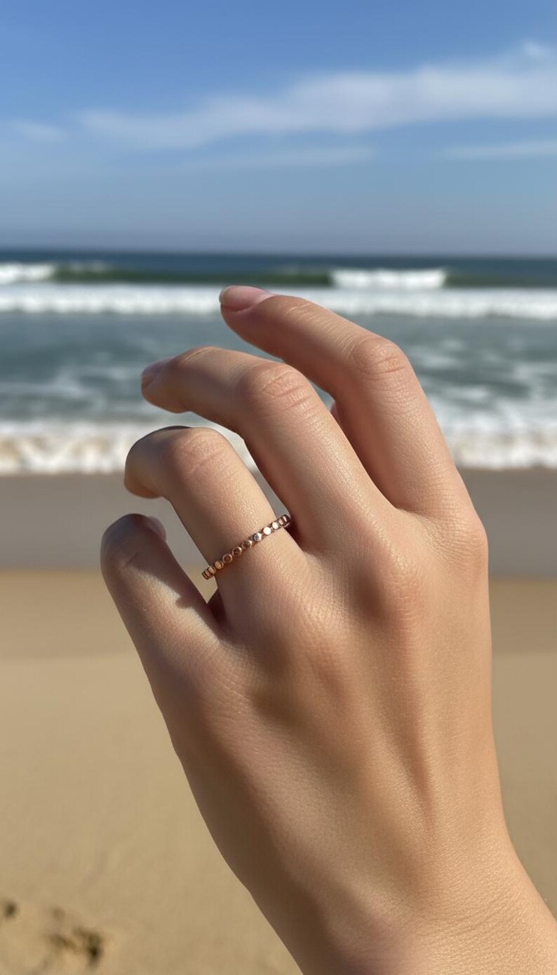 Puede incluir: Un anillo de oro rosa con un delicado dise&ntilde;o de cuentas. El anillo se muestra en una mano con un fondo de playa y oc&eacute;ano. El dise&ntilde;o del anillo presenta peque&ntilde;as cuentas espaciadas uniformemente alrededor de la banda.