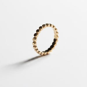 Puede incluir: Un anillo dorado con un dise&ntilde;o de puntos repetidos. El anillo est&aacute; sobre un fondo blanco, con una sombra proyectada hacia la izquierda. El dise&ntilde;o del anillo presenta peque&ntilde;os puntos espaciados uniformemente alrededor de la banda.