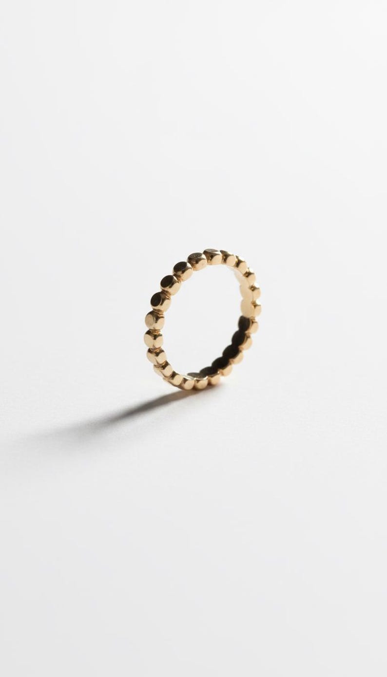 Puede incluir: Un anillo dorado con un dise&ntilde;o de puntos repetidos. El anillo est&aacute; sobre un fondo blanco, con una sombra proyectada hacia la izquierda. El dise&ntilde;o del anillo presenta peque&ntilde;os puntos espaciados uniformemente alrededor de la banda.