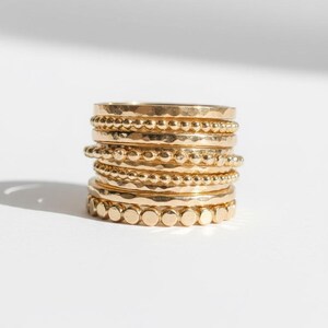Puede incluir: Una pila de anillos dorados con diversas texturas y dise&ntilde;os. Los anillos incluyen bandas martilladas, con cuentas y lisas, creando un efecto en capas. Los anillos se exhiben sobre una superficie blanca, resaltando su brillo met&aacute;lico.