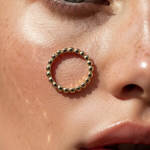 Puede incluir: Primer plano de un anillo dorado con dise&ntilde;o de cuentas. El anillo est&aacute; colocado en la cara de una persona. La persona tiene piel clara, pecas y ojos verdes.