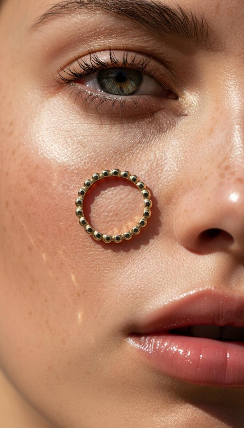 Puede incluir: Primer plano de un anillo dorado con dise&ntilde;o de cuentas. El anillo est&aacute; colocado en la cara de una persona. La persona tiene piel clara, pecas y ojos verdes.