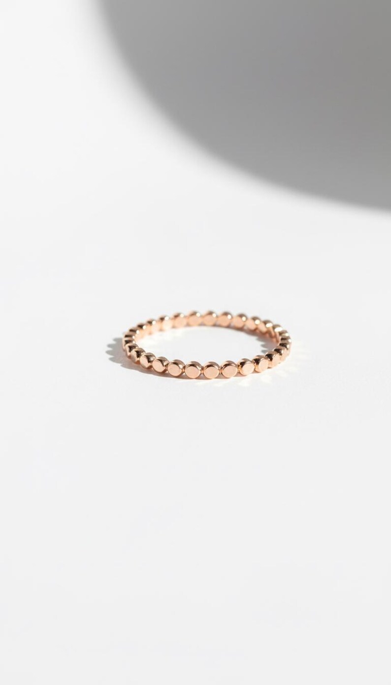 Puede incluir: Un delicado anillo de oro rosa con una serie de peque&ntilde;os acentos circulares espaciados uniformemente. El anillo se encuentra sobre un fondo blanco limpio, resaltando su simple elegancia. El dise&ntilde;o del anillo es minimalista y moderno.