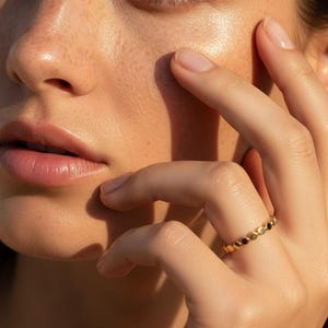 Puede incluir: Primer plano de un rostro y una mano. Un anillo de oro con peque&ntilde;os detalles circulares adorna un dedo. La piel tiene un tono c&aacute;lido, con pecas visibles y un brillo suave y natural. La atenci&oacute;n se centra en el anillo y la piel.