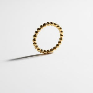 Puede incluir: Un anillo dorado compuesto por peque&ntilde;as esferas espaciadas uniformemente. El anillo est&aacute; sobre un fondo blanco, proyectando una sombra sutil. La joya es simple y elegante.