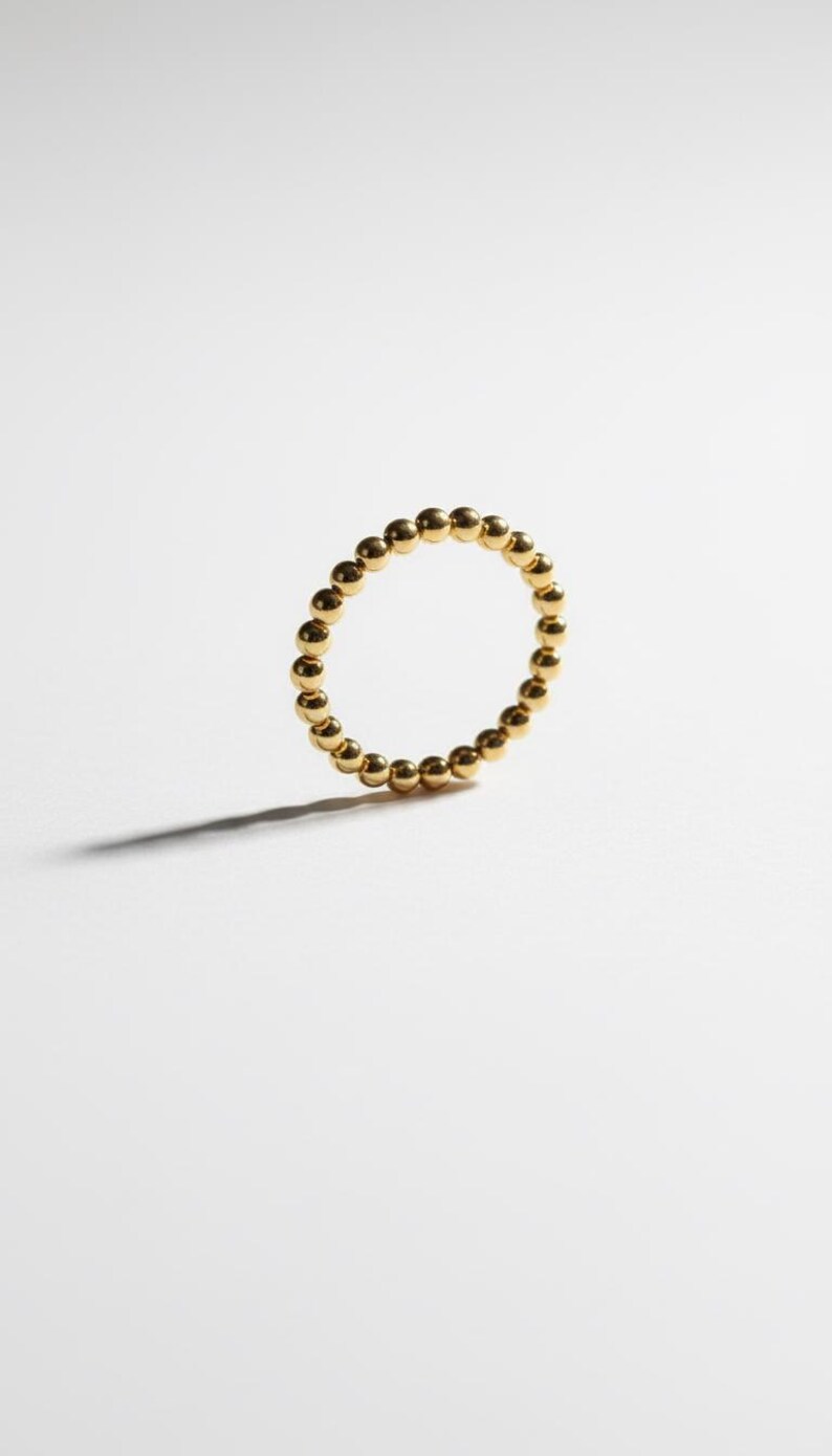 Puede incluir: Un anillo dorado compuesto por peque&ntilde;as esferas espaciadas uniformemente. El anillo est&aacute; sobre un fondo blanco, proyectando una sombra sutil. La joya es simple y elegante.