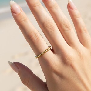 Puede incluir: Un anillo dorado con un dise&ntilde;o de cuentas se lleva en un dedo. El anillo est&aacute; hecho de peque&ntilde;as cuentas redondas que est&aacute;n unidas para formar una banda. El fondo es una escena de playa con arena y agua.