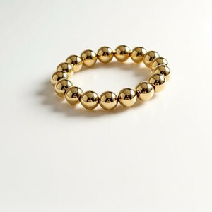 Puede incluir: Una pulsera dorada con cuentas y dise&ntilde;o el&aacute;stico. La pulsera presenta m&uacute;ltiples cuentas redondas y brillantes de color dorado. La joya se exhibe sobre un fondo blanco, resaltando el color y el dise&ntilde;o de la pulsera. Este accesorio es adecuado para el uso diario.