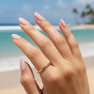 Puede incluir: Un anillo de oro con superficie texturizada se lleva en una mano con u&ntilde;as cuidadas. El anillo se encuentra sobre un fondo de playa con cielo azul y oc&eacute;ano.