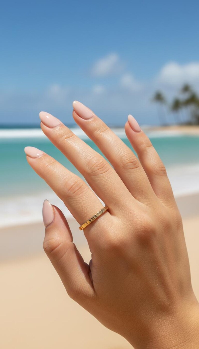 Puede incluir: Un anillo de oro con superficie texturizada se lleva en una mano con u&ntilde;as cuidadas. El anillo se encuentra sobre un fondo de playa con cielo azul y oc&eacute;ano.