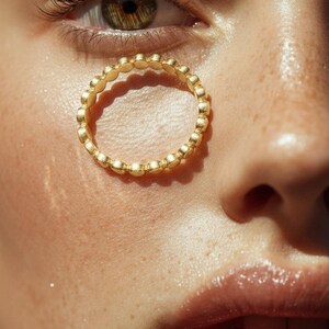 Puede incluir: Primer plano de un anillo dorado con un dise&ntilde;o de cuentas. El anillo est&aacute; posicionado cerca de un ojo con tonos verdes y marrones, y la piel tiene una apariencia natural y h&uacute;meda. Los detalles del anillo son claramente visibles.