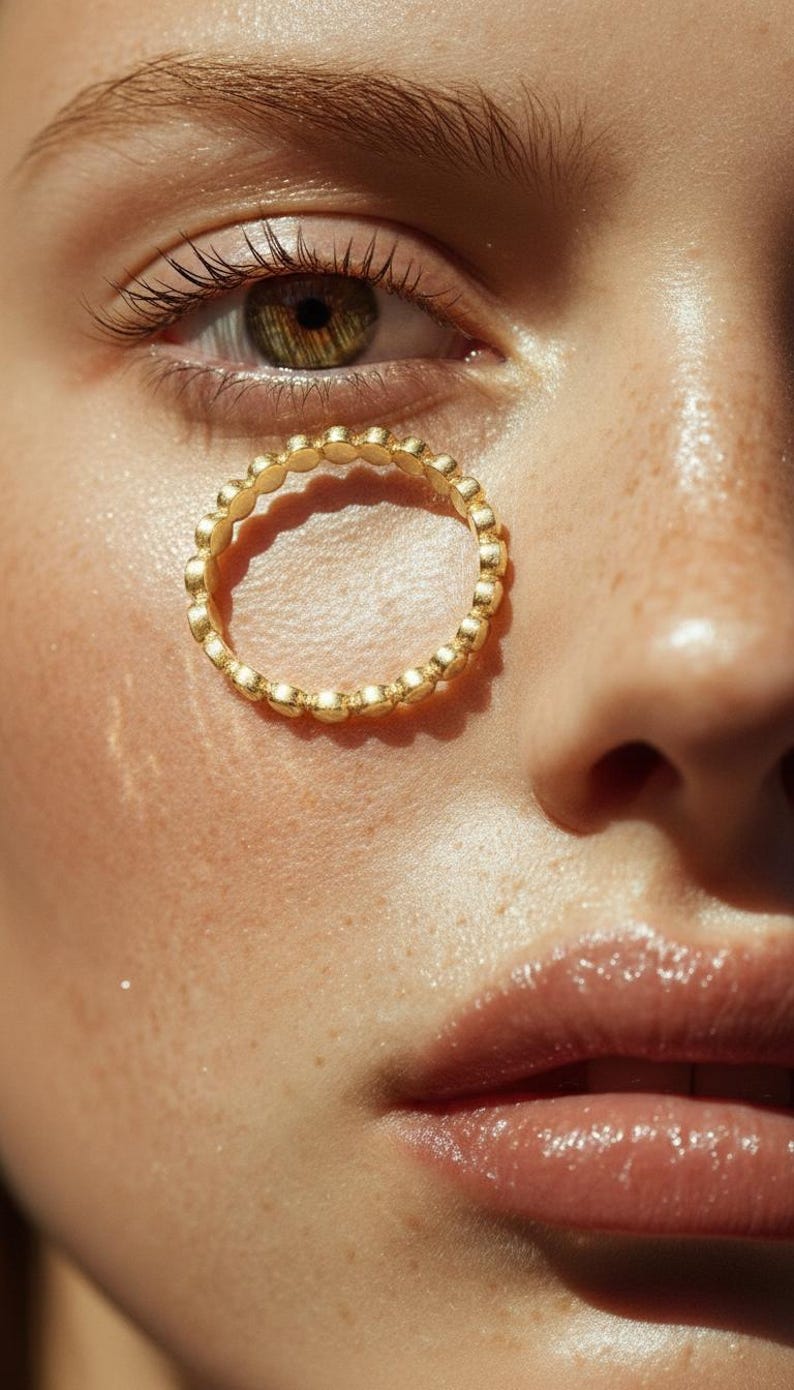 Puede incluir: Primer plano de un anillo dorado con un dise&ntilde;o de cuentas. El anillo est&aacute; posicionado cerca de un ojo con tonos verdes y marrones, y la piel tiene una apariencia natural y h&uacute;meda. Los detalles del anillo son claramente visibles.