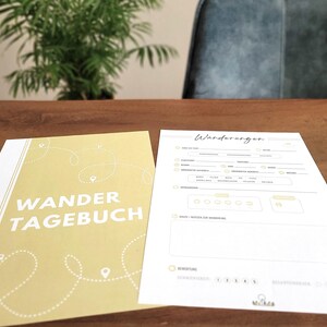 Może przedstawiać: Na drewnianej powierzchni leżą dziennik i pusta strona. Okładka dziennika jest jasnożółta z białymi słowami "WANDER TAGEBUCH". Pusta strona ma u góry słowo "Wanderungen". W tle zielona roślina.