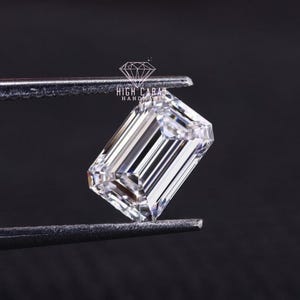 Op de afbeelding: Een heldere diamant met smaragd-slijpsel wordt door een pincet tegen een zwarte achtergrond gehouden. De diamant heeft een rechthoekige vorm met trapsgewijze facetten, wat een briljante glans creëert. De tekst "HIGH CARAT HANDMADE" is zichtbaar.