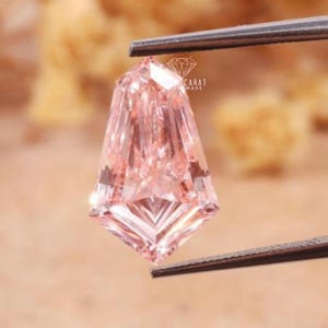Può includere: Una pietra preziosa rosa chiaro con taglio a scudo tenuta da pinzette in metallo. La pietra presenta una superficie sfaccettata con forma geometrica e base appuntita. Un piccolo logo con il testo "AURA CARAT HANDMADE" è visibile sulla pietra.