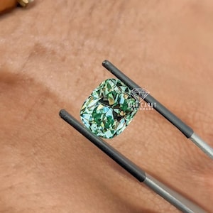 May include: A cushion-cut gemstone with a vibrant green hue and multifaceted surface held by metal tweezers. The stone features a clear, reflective quality with visible internal light refraction. The text HIGH CARAT HANDMADE is visible.