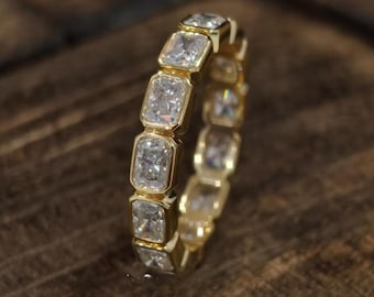 May include: A gold eternity band featuring a continuous row of rectangular radiant-cut clear stones set in individual bezel settings. The ring displays a polished metallic finish and a uniform arrangement of faceted gemstones encircling the entire band.