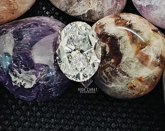 May include: A clear, oval-cut gemstone with multiple facets reflecting light, positioned among several smooth, polished stones in shades of purple, white, and brown. Below the gemstone, the text reads HIGH CARAT HANDMADE with a diamond icon.