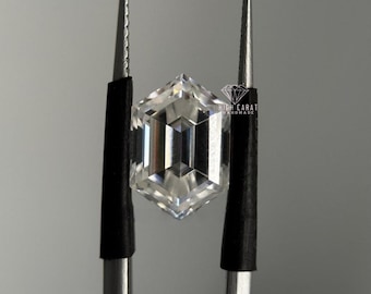 May include: A clear, colorless, hexagonal-cut gemstone held by metal tweezers. The stone features a geometric, multi-faceted surface that reflects light. A small logo with the text HIGH CARAT HANDMADE is visible near the top right edge.