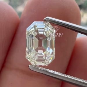 Pode incluir: Uma pedra preciosa transparente de corte esmeralda com uma superfície facetada que reflete a luz, segurada por pinças de metal. A pedra apresenta um logótipo em forma de diamante e o texto HIGH CARAT HANDMADE impresso a branco.
