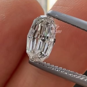 Op de afbeelding: Een heldere, rechthoekig geslepen diamant, vastgehouden door een pincet. De diamant heeft meerdere facetten die licht reflecteren. De tekst "HIGH CARAT HANDMADE" is zichtbaar op de achtergrond.