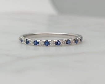 May include: A thin silver-toned metal band ring featuring an alternating pattern of small, round, clear white stones and deep blue sapphire-colored stones set along the top half of the band in a continuous, delicate, and polished metal setting.