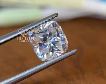 May include: A cushion-cut, clear, faceted gemstone held by metal tweezers. The stone displays high brilliance and light refraction. The text HIGH CARAT HANDMADE with a diamond icon is printed in white on the left side of the gemstone surface.