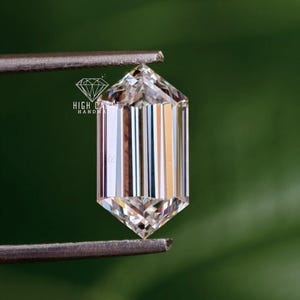 Pode incluir: Uma pedra preciosa de corte hexagonal com uma superfície facetada clara que exibe reflexos verticais e refrações internas. A pedra é segurada por uma pinça metálica e apresenta o texto HIGH CARAT HANDMADE impresso em branco.