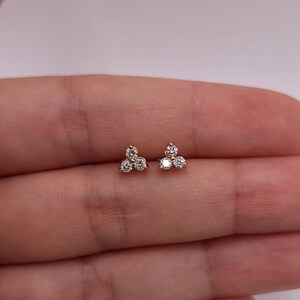 May include: A pair of gold stud earrings, each with three clear, round gemstones arranged in a cluster. The earrings are presented against a plain background, emphasizing their delicate design and sparkle.
