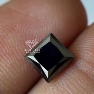 Op de afbeelding: Een vierkant geslepen, zwarte edelsteen met een gefacetteerd oppervlak. De edelsteen is geplaatst tegen een vage achtergrond. De tekst "HIGH CARAT HANDMADE" wordt in het wit weergegeven, met een diamantafbeelding boven de tekst.