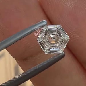Op de afbeelding: Een heldere, kleurloze, zeshoekig geslepen edelsteen met een gefacetteerd oppervlak. De steen heeft een geometrisch, meerlaags intern patroon. De tekst "HIGH CARAT HANDMADE" en een diamanten icoon zijn in het wit op de steen gedrukt.