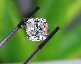 May include: A cushion-cut, colorless gemstone held by black metal tweezers. The stone features a brilliant, multifaceted surface that reflects light. A white logo with the text HIGH CARAT HANDMADE is visible on the surface of the gemstone.