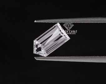 May include: A clear, colorless, baguette-cut gemstone held by metal tweezers. The stone features a rectangular shape with elongated facets that reflect light. A small logo with the text HIGH CARAT HANDMADE is visible near the stone.