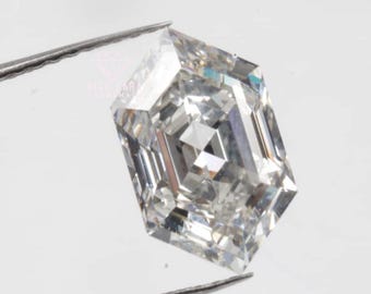 May include: A clear, colorless, hexagonal-cut gemstone held by metal tweezers. The stone features a geometric, multi-faceted surface that reflects light, creating a brilliant, sparkling appearance. The text HIGH CARAT HANDMADE is visible.