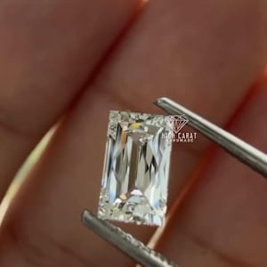 Op de afbeelding: Een rechthoekig geslepen, heldere diamant, vastgehouden door een pincet. De diamant heeft vele facetten die het licht reflecteren. De tekst "HIGH CARAT HANDMADE" is zichtbaar op de achtergrond.