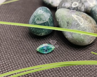 May include: A marquise-cut teal gemstone with a faceted surface, positioned near several smooth, rounded green moss agate stones. The gemstone features a small white logo with the text HIGH CARAT HANDMADE printed above it.