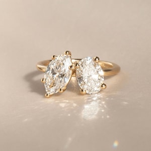 May include: A gold ring featuring two large, sparkling diamonds. One diamond is a marquise cut, and the other is a pear cut. The ring has a simple, elegant design, with the diamonds set close together on the band.