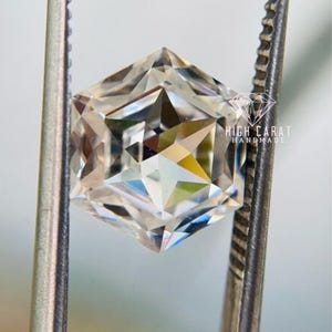 May include: A clear, colorless, hexagonal-cut gemstone held by metal tweezers. The stone features intricate, sharp facets that reflect light, creating a star-like pattern. The text HIGH CARAT HANDMADE is printed in white on the right side of the stone.