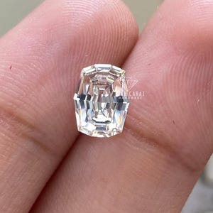 May include: A clear, colourless gemstone with an emerald-style step cut, featuring multiple concentric rectangular facets that create a bright, reflective surface. The stone is marked with a small logo and the text AH CARAT HANDMADE.