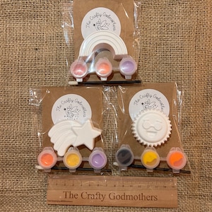 May include: Three paint-your-own craft kits featuring a rainbow, a shooting star, and a sun with a smiley face. Each kit includes a white ceramic piece, paint pots in various colors, and a paintbrush.