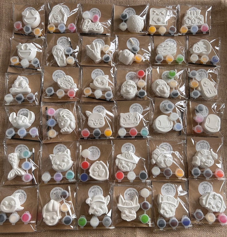 May include: A set of 20 paint your own ceramic ornaments, each packaged individually with a small pot of paint. The ornaments feature various designs, including cartoon characters, animals, and abstract shapes.