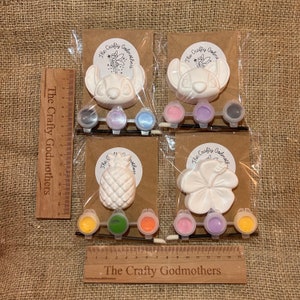 May include: Four paint-your-own ceramic ornaments in individual clear plastic bags. The ornaments are a pineapple, a hibiscus flower, and two Stitch characters from Lilo & Stitch. Each bag includes a small paintbrush and a set of acrylic paints in various colors. The bags are labeled "The Crafty Godmothers".