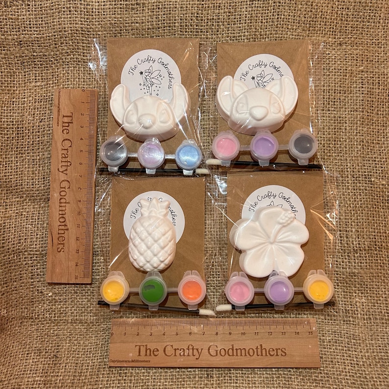 May include: Four paint-your-own ceramic ornaments in individual clear plastic bags. The ornaments are a pineapple, a hibiscus flower, and two Stitch characters from Lilo & Stitch. Each bag includes a small paintbrush and a set of acrylic paints in various colors. The bags are labeled "The Crafty Godmothers".
