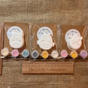 May include: Three paint-your-own ceramic figurines, a cat, a bird, and a unicorn, with small pots of paint in various colors. The text "The Crafty Godmothers" is on the brown paper backing and on a wooden ruler.