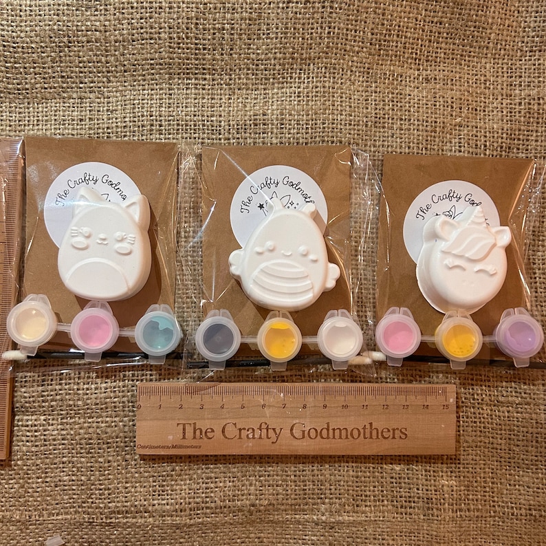 May include: Three paint-your-own ceramic figurines, a cat, a bird, and a unicorn, with small pots of paint in various colors. The text "The Crafty Godmothers" is on the brown paper backing and on a wooden ruler.