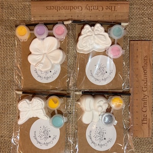 May include: Four paint-your-own ceramic craft kits with different designs: a butterfly, a bee, a flower, and another butterfly. Each kit includes a small paintbrush, a small ceramic piece, and four small pots of paint.