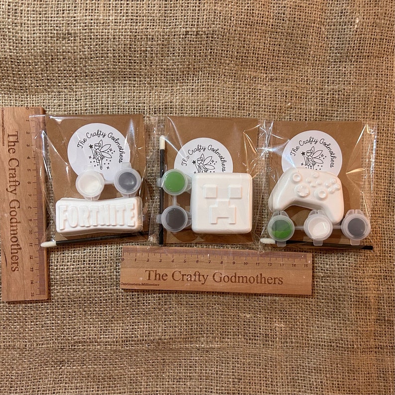 May include: Three paint-your-own kits featuring popular video game themes. The kits include a white ceramic figurine, paint pots, a paintbrush, and a small container of water. The figurines are shaped like a controller, a Minecraft block, and the word "FORTNITE".